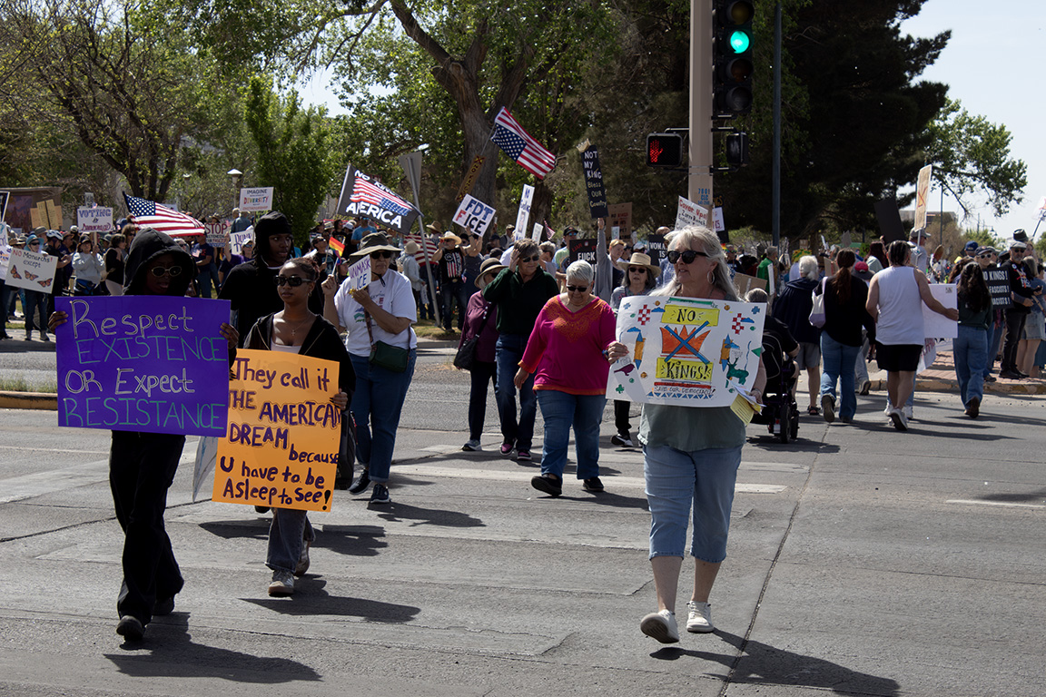 Protesters cross Picacho Avenue holding signs that read "Respect Existence or Expect Resistance," "They call it the AMERICAN DREAM because u have to be Asleep to see!" and "No Kings!" March 28, 2026.