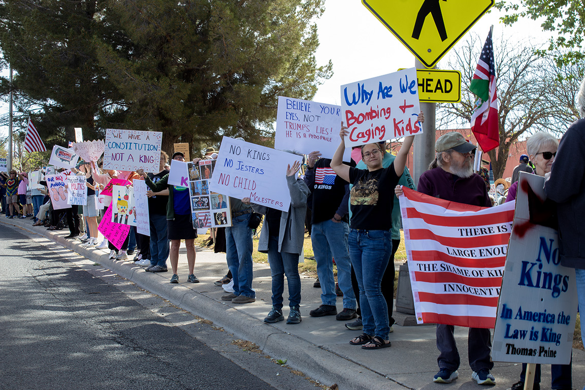 Dozens of protesters stand along Picacho Street with signs that read "We have a Constitution not a king" and "Why are we bombing and caging kids?" March 28, 2026.